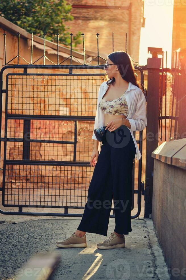 Portrait of a girl in dark sunglasses posing in city. Dressed in top with floral print, white shirt, black trousers, waist bag and sneakers. photo
