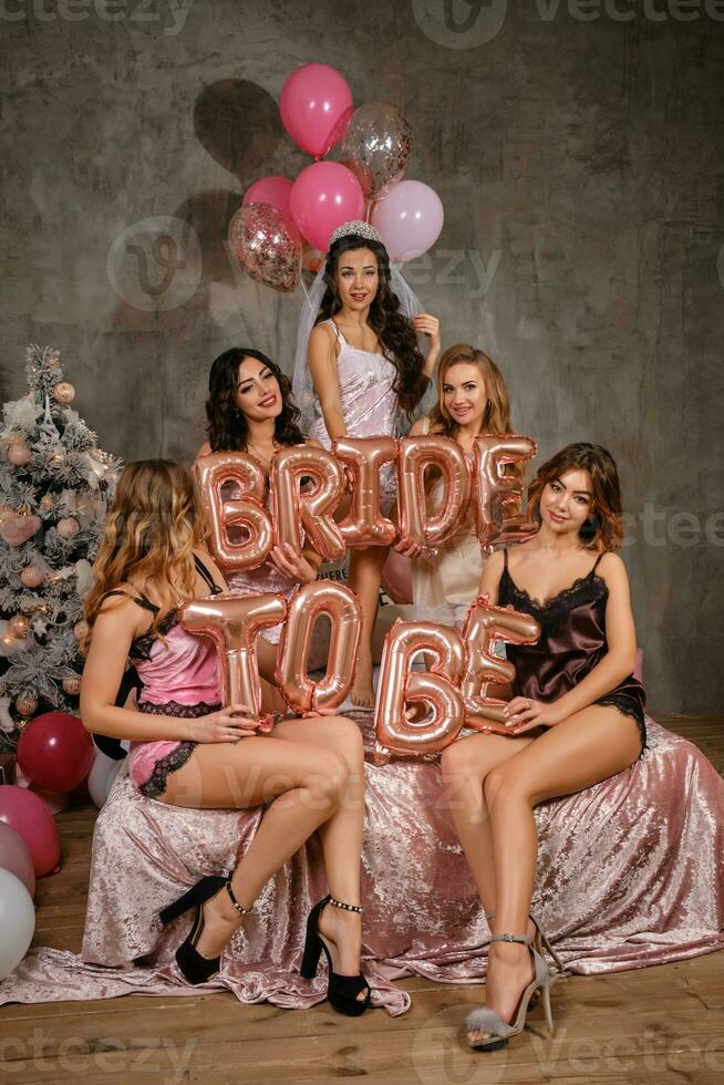 Ladies in sexy lingerie and bride in veil, enjoying a hen-party, sitting on bed, holding balloons, smiling. New year tree, decorations. Close-up. photo