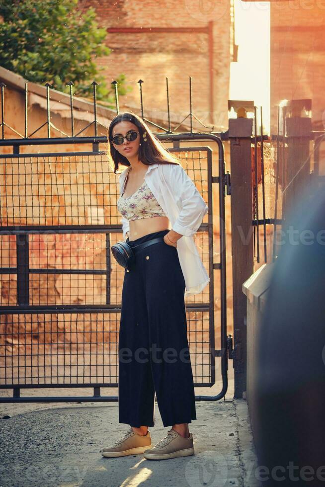 Portrait of a girl in dark sunglasses posing in city. Dressed in top with floral print, white shirt, black trousers, waist bag and sneakers. photo