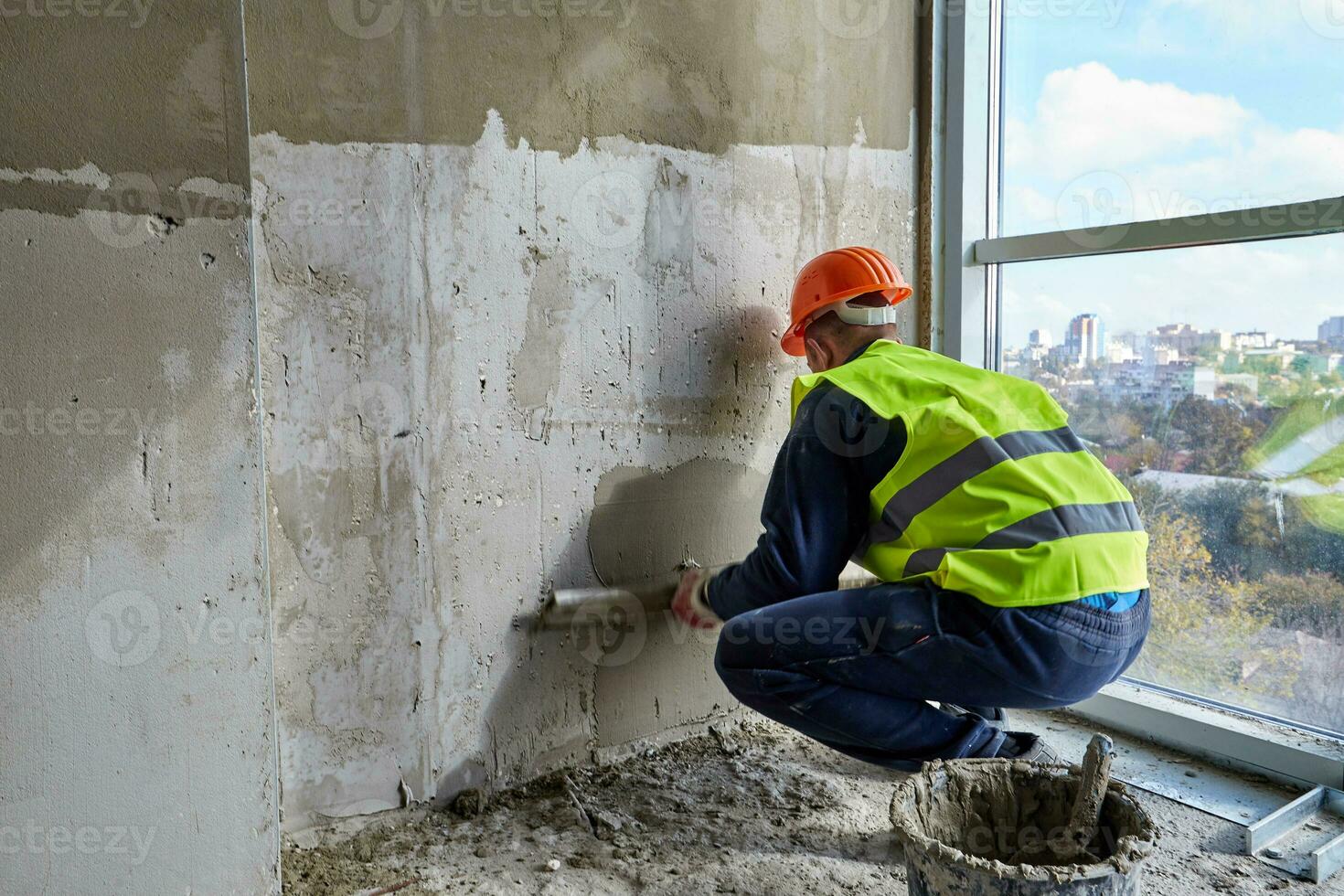 experto trabajador en trabajando ropa y naranja difícil sombrero es poniendo masilla en el muro, utilizando espátula. plano de un edificio debajo construcción foto