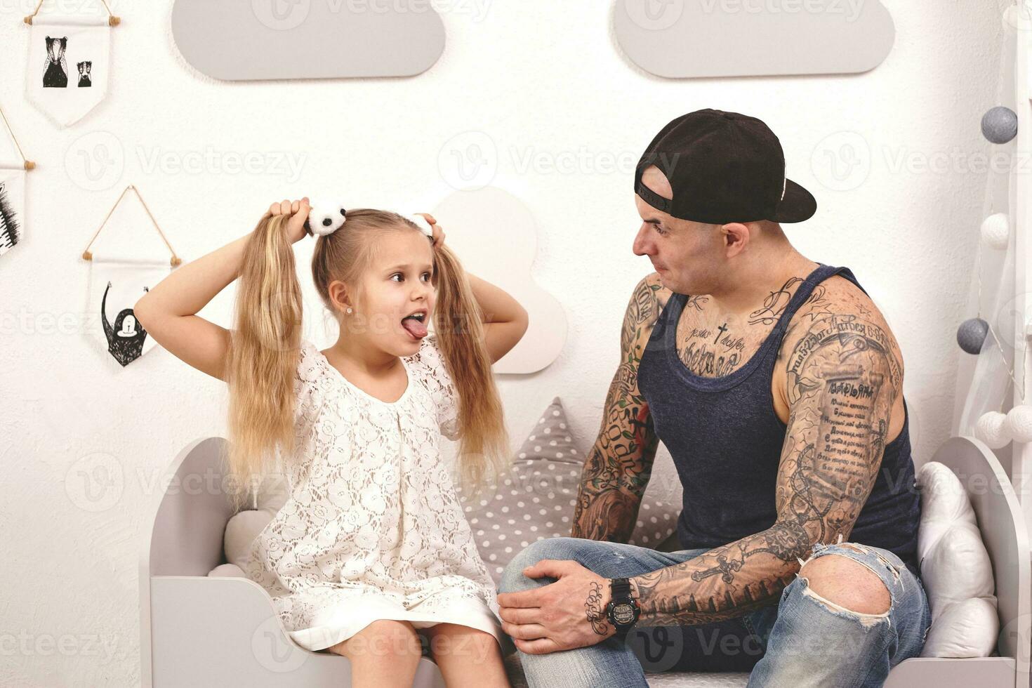 Funny time Tattoed father in a cap and his child are playing at home. Dad is doing his daughter's hair in her bedroom. Family holiday and togetherness photo