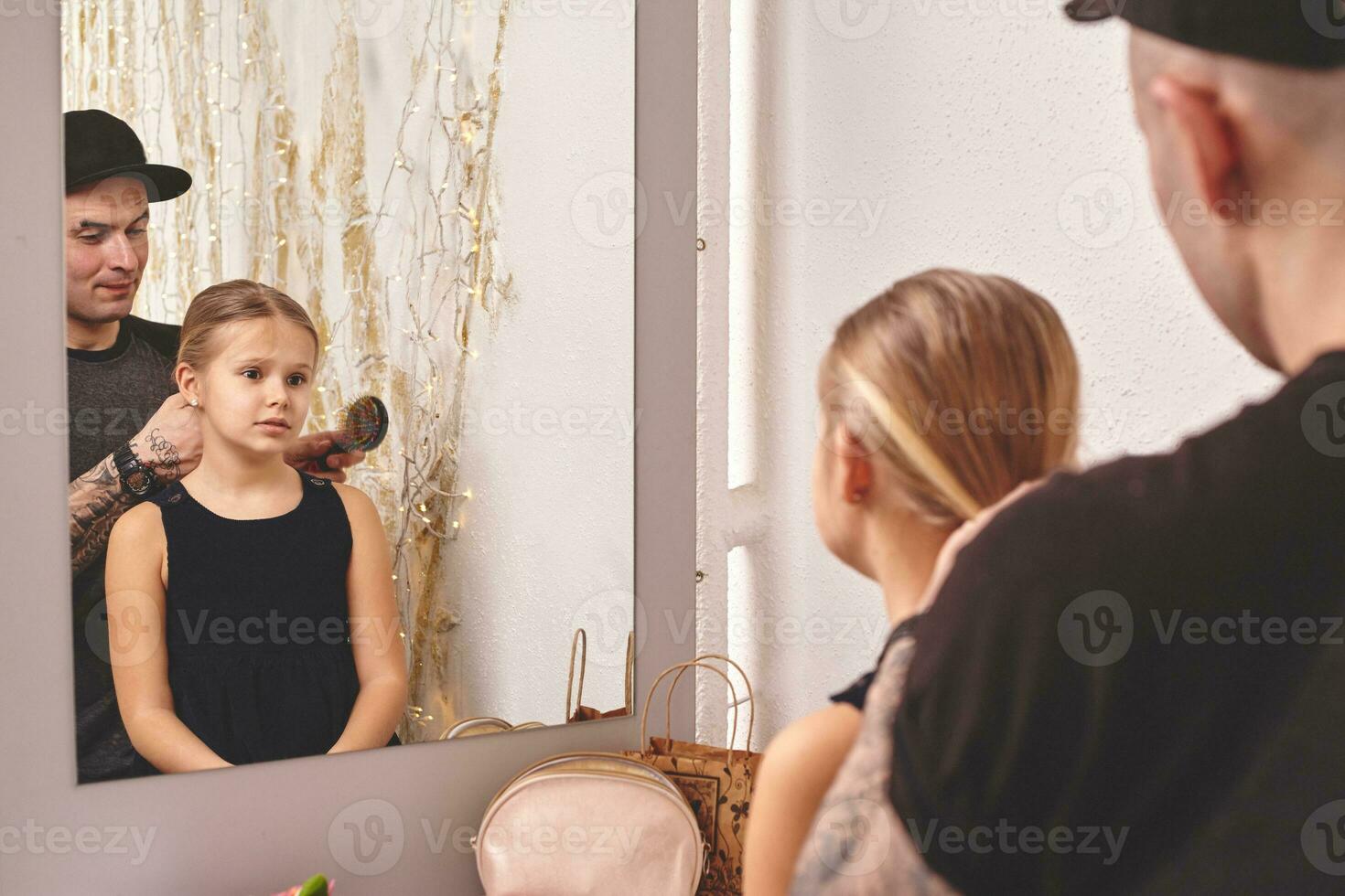 Cute little daughter and her tattoed dad are playing together near a mirror. Dad is doing his daughter's hair. Family holiday and togetherness. photo