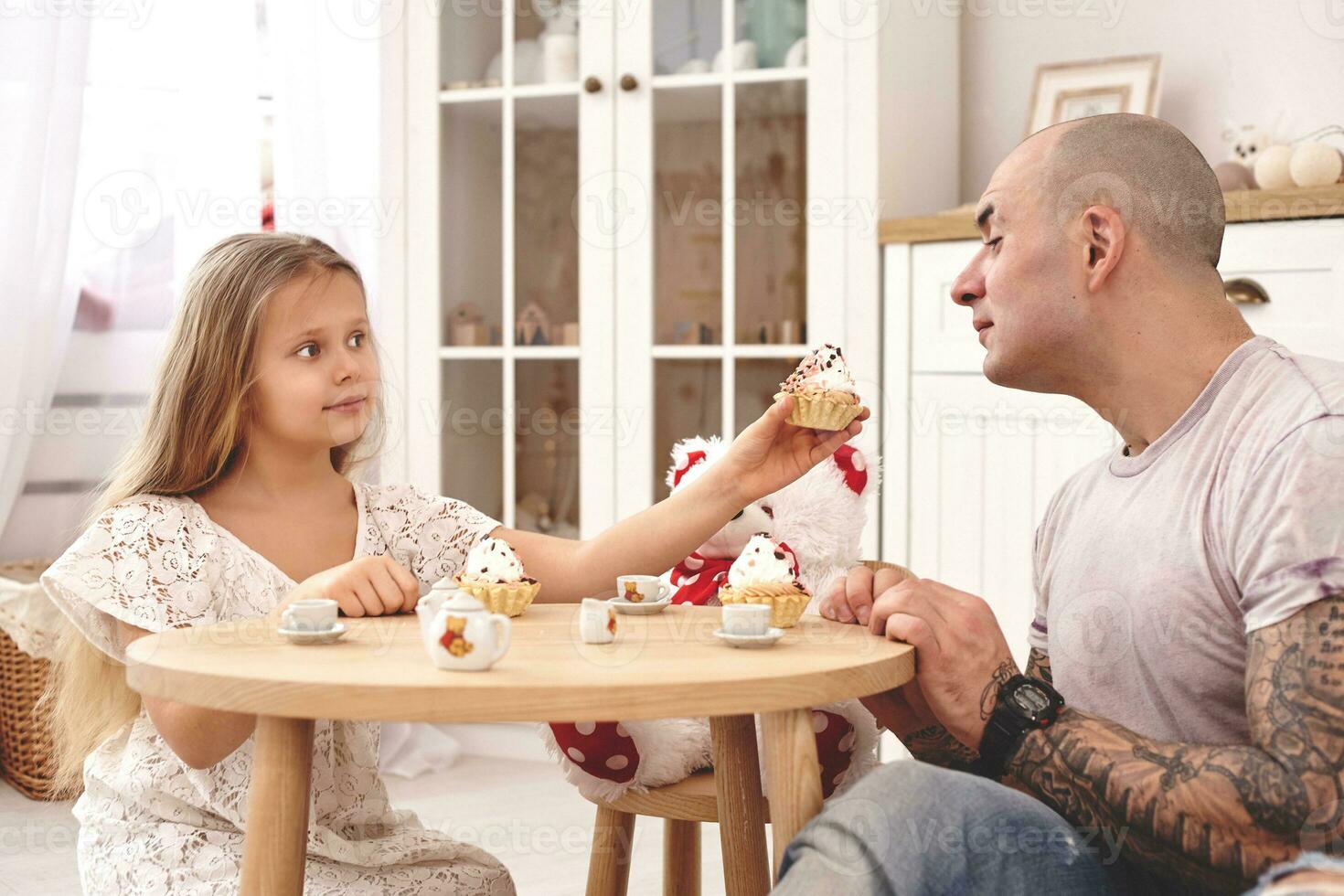 adorable hija vistiendo un blanco vestir con su amoroso padre. ellos son Bebiendo té desde un juguete platos en un moderno niño habitación. contento familia. foto