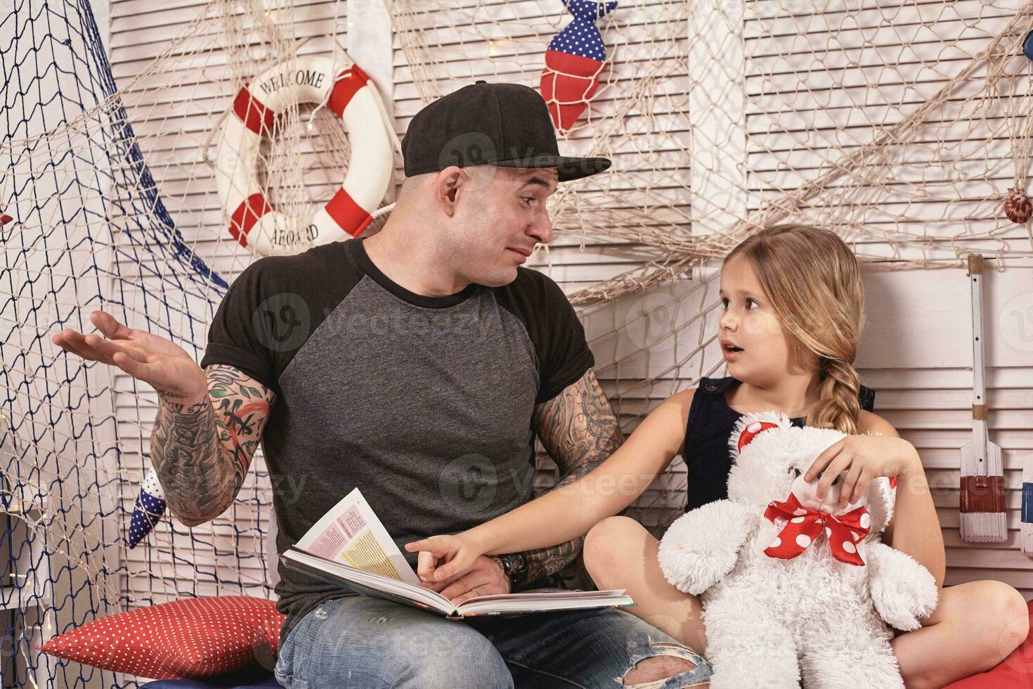 Handsome tattoed man is spending time with his little cute daughter. Reading fairytales while daughter is sitting nearby. Happy family. photo