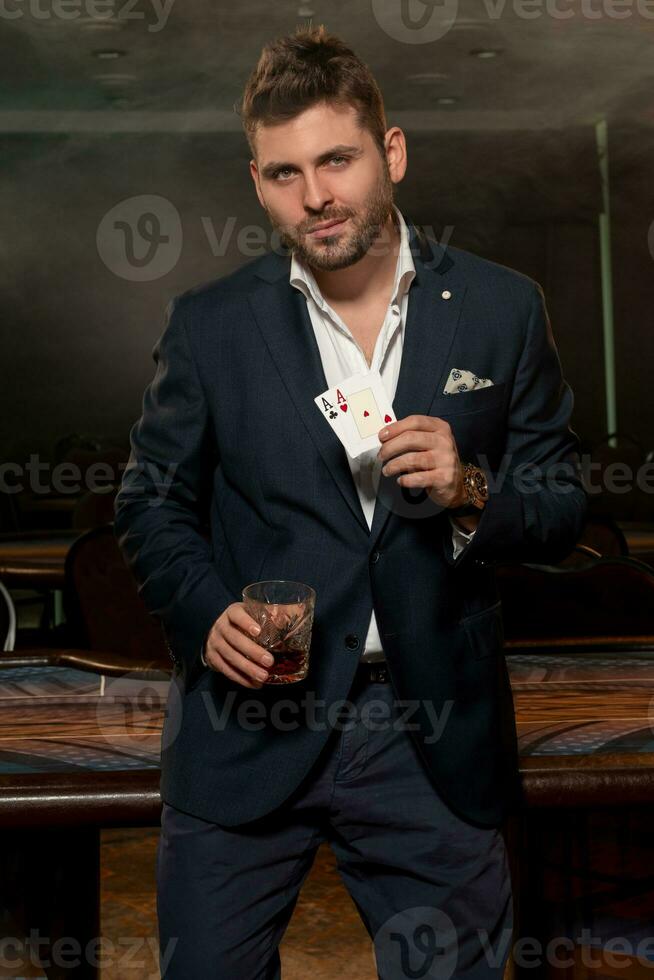 Successful young gambler with glass of drink and pair of aces near gaming table in casino photo
