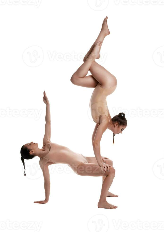 dos flexible muchachas gimnastas en beige leotardos son ejecutando ejercicios al revés abajo utilizando apoyo y posando aislado en blanco antecedentes. de cerca. foto