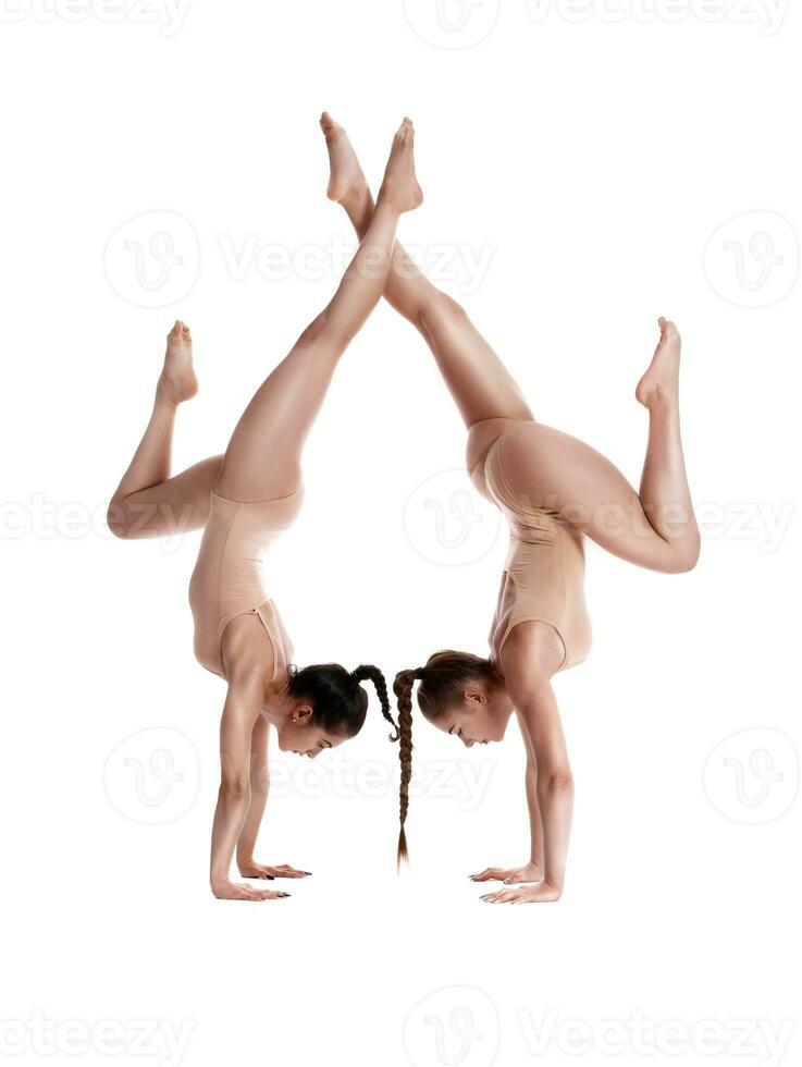 dos flexible muchachas gimnastas en beige leotardos son ejecutando ejercicios al revés abajo utilizando apoyo y posando aislado en blanco antecedentes. de cerca. foto