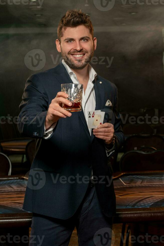 sonriente chico con dos ases en mano levantamiento vaso de whisky a exitoso póker juego foto