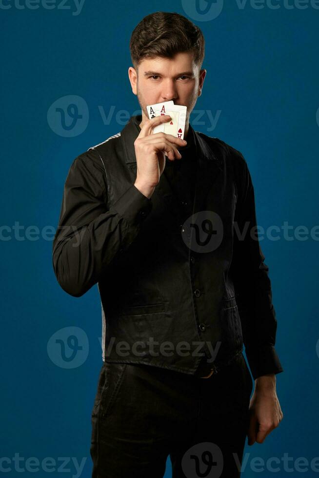 Noob in poker, in black vest, trousers and shirt. Holding two playing cards while posing against blue studio background. Gambling, casino. Close-up. photo