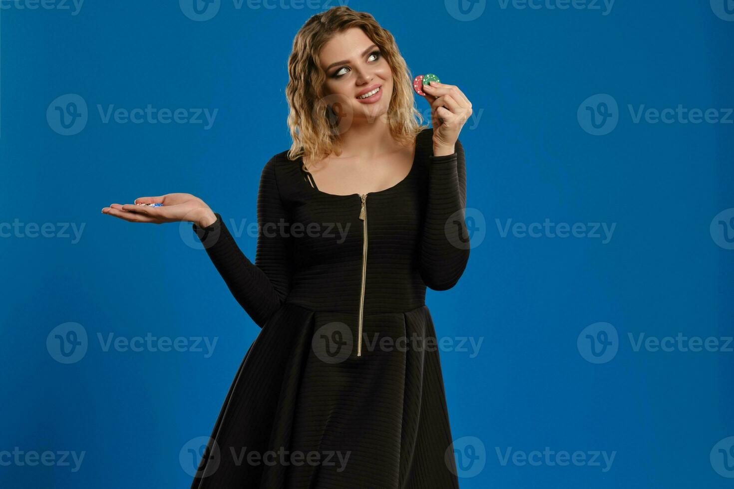 Blonde female in black dress is smiling, showing some colorful chips, posing against blue background. Gambling entertainment, poker, casino. Close-up. photo