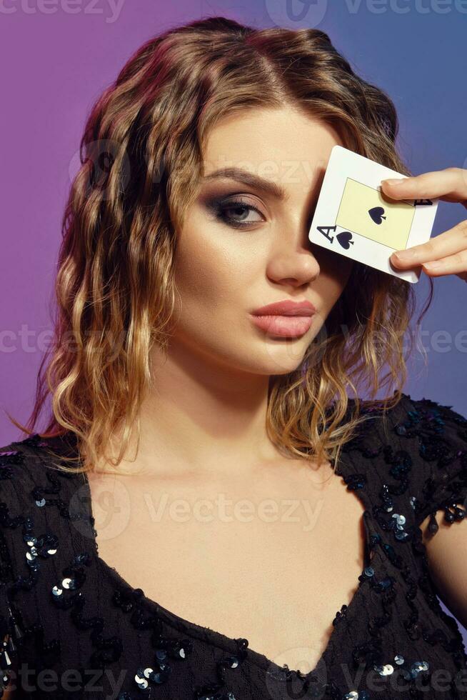 Blonde lady in black sequin dress has covered her eye with ace of spades, posing on colorful studio background. Gambling, poker, casino. Close-up. photo