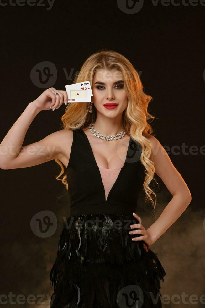 Blonde woman in black dress with feathers and necklace. Smiling, showing two playing cards, posing on black smoky background. Poker, casino. Close-up photo