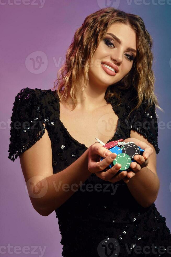 Blonde female in black sequin dress is smiling, showing handful of multicolored chips, posing on colorful studio background. Poker, casino. Close-up. photo