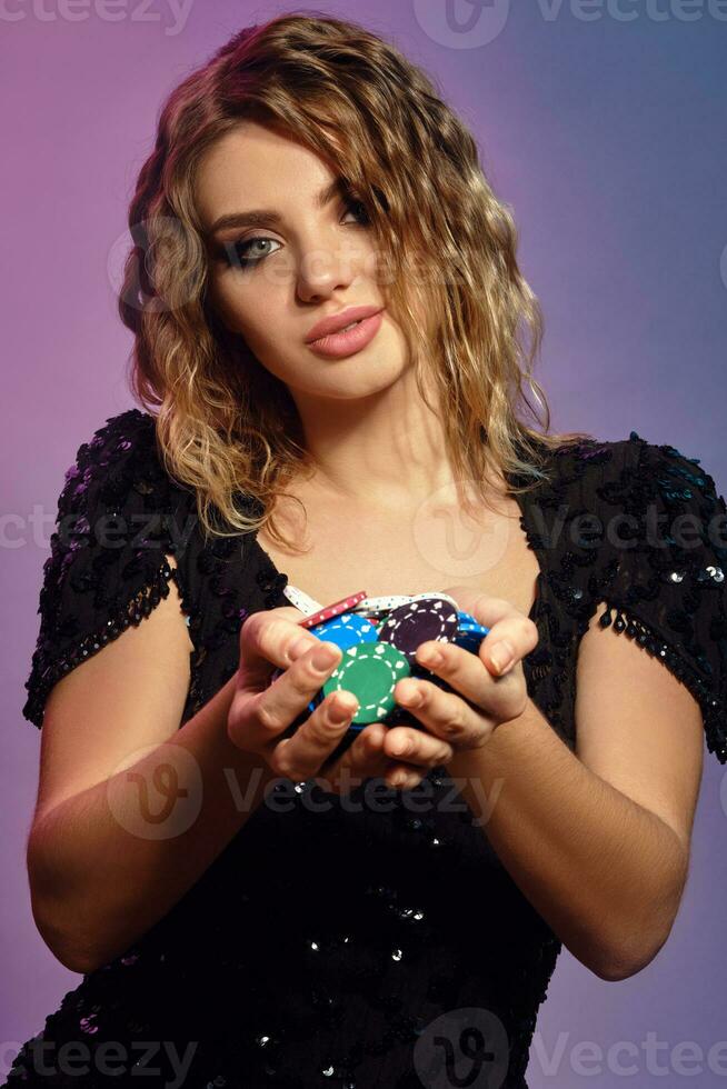 Blonde lady in black sequin dress is holding handful of multicolored chips, posing on colorful studio background. Gambling, poker, casino. Close-up. photo