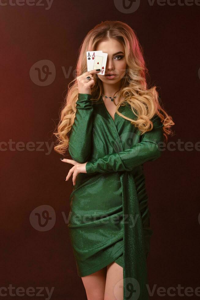 Blonde curly woman in green dress and jewelry. She covered half of her face with two aces, posing on brown background. Poker, casino. Close-up photo