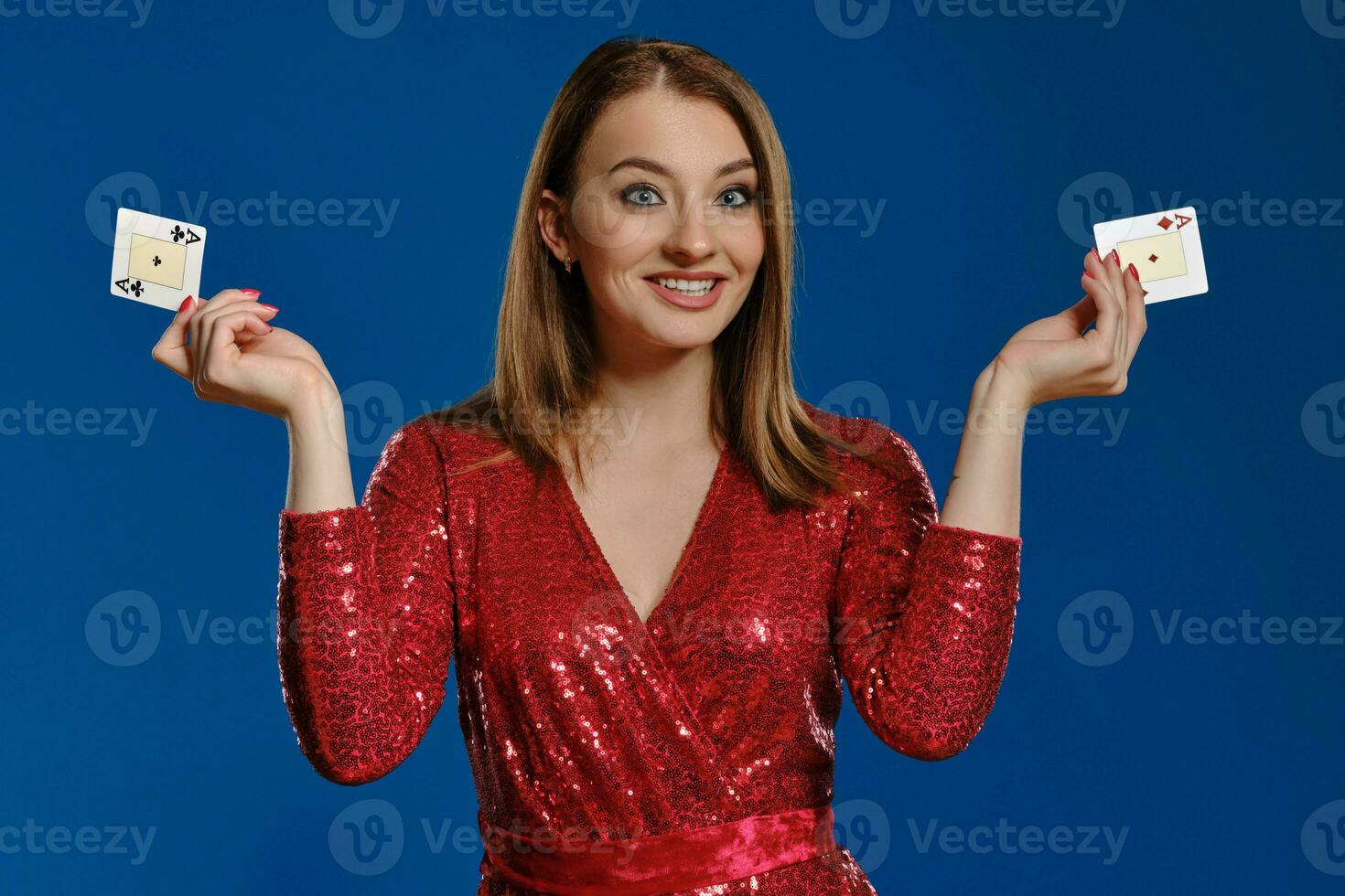 Blonde lady with make-up, in red sequin dress is smiling, showing two aces, posing on blue background. Gambling, poker, casino. Close-up. photo