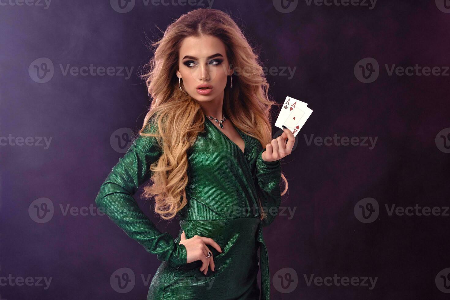 Blonde woman in green stylish dress and jewelry. Showing two playing cards, hand on waist, posing on purple smoky background. Poker, casino. Close-up photo