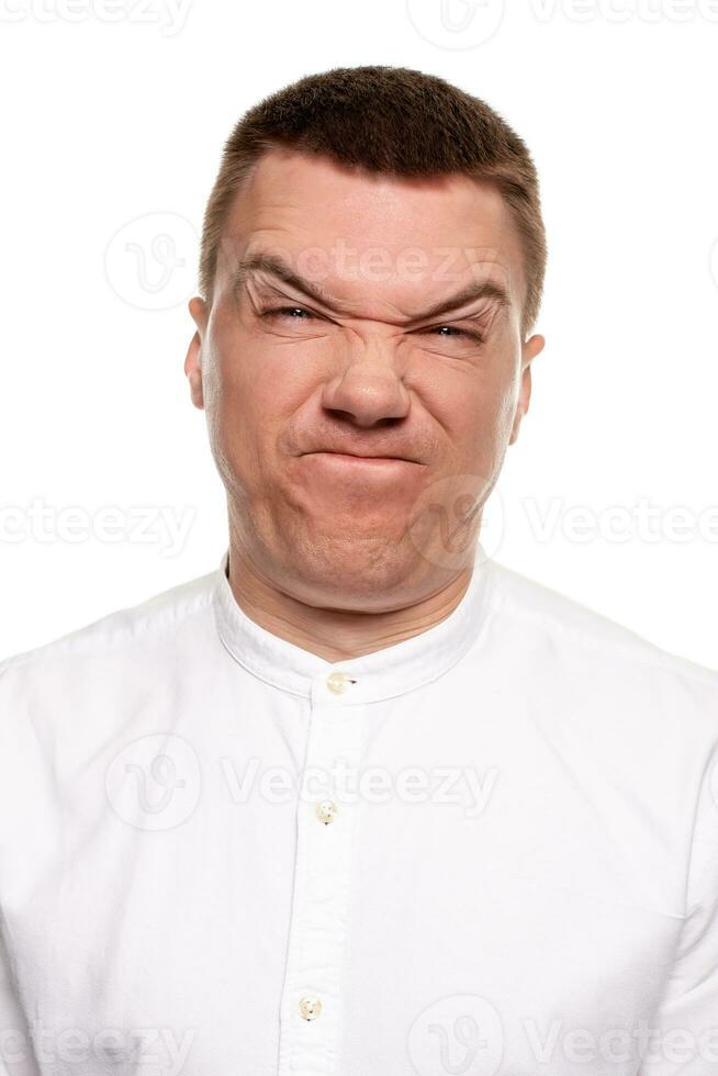 Charming handsome young man in a white shirt is making faces, while standing isolated on a white background photo