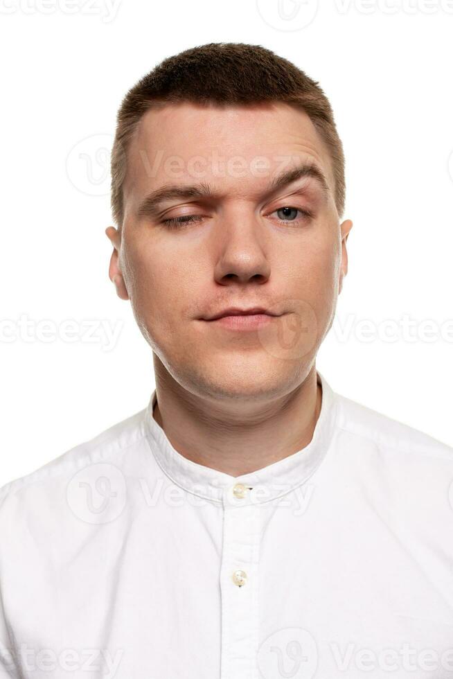 Charming handsome young man in a white shirt is making faces, while standing isolated on a white background photo