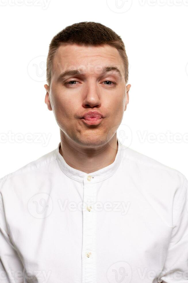 Charming handsome young man in a white shirt is making faces, while standing isolated on a white background photo