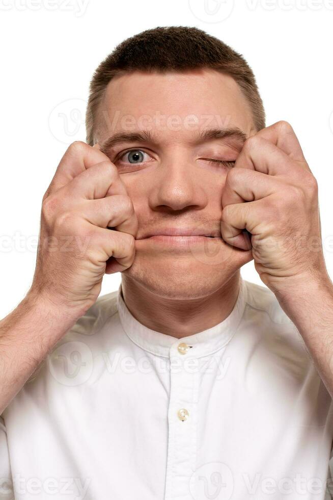 Charming handsome young man in a white shirt is making faces, while standing isolated on a white background photo