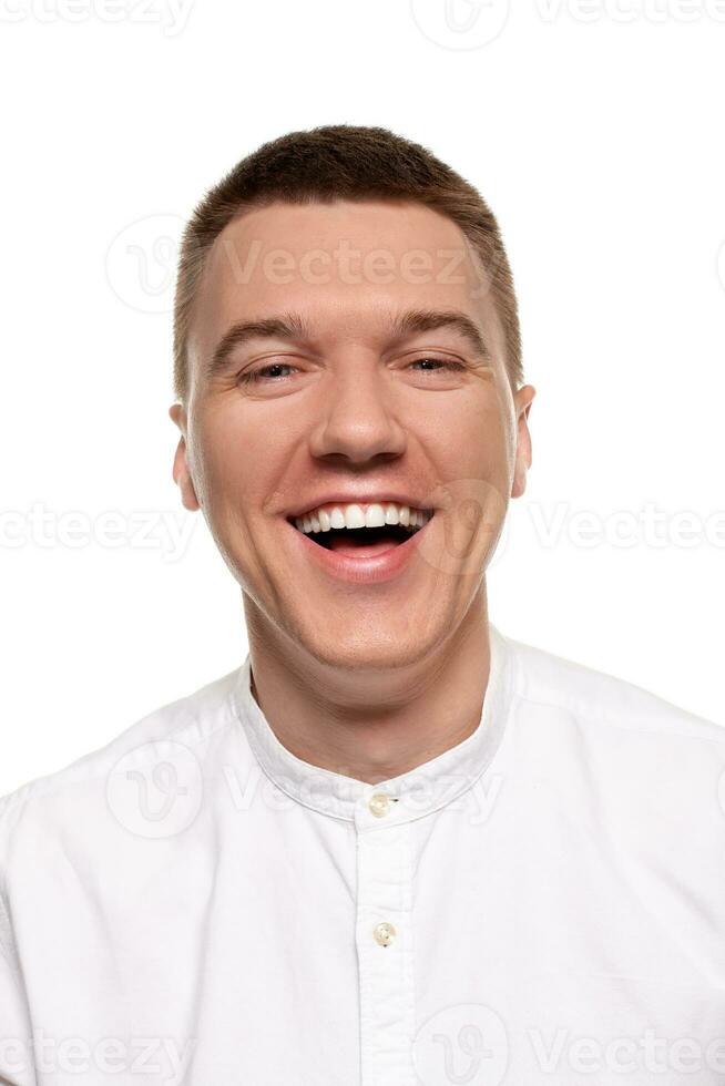 Charming handsome young man in a white shirt is making faces, while standing isolated on a white background photo