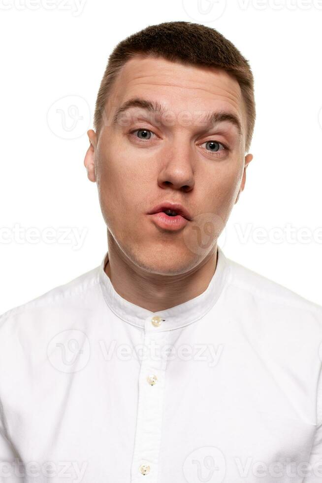 encantador hermoso joven hombre en un blanco camisa es haciendo caras, mientras en pie aislado en un blanco antecedentes foto