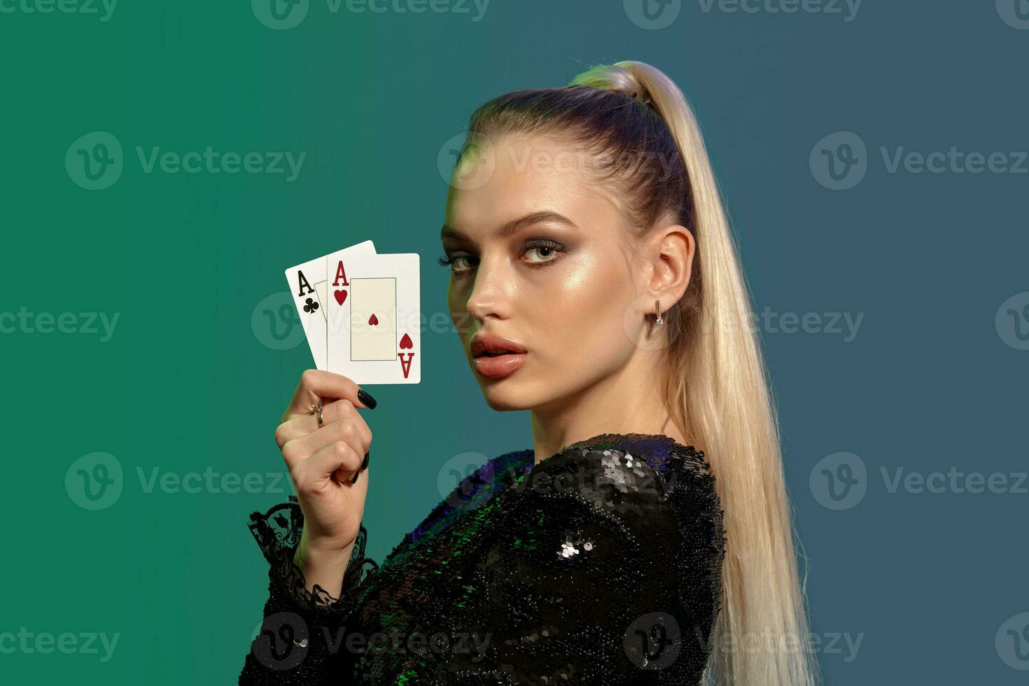 Blonde model with ponytail, in black sequin dress. Showing two aces, posing sideways on colorful background. Gambling, poker, casino. Close-up photo