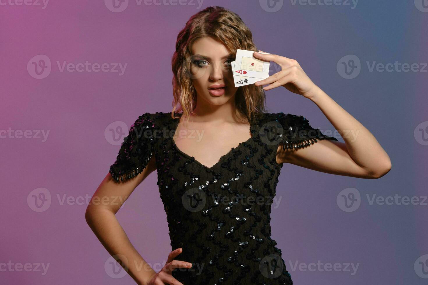 Brown-haired lady in black shiny dress has covered eye with two playing cards, posing on colorful background. Gambling, poker, casino. Close-up. photo