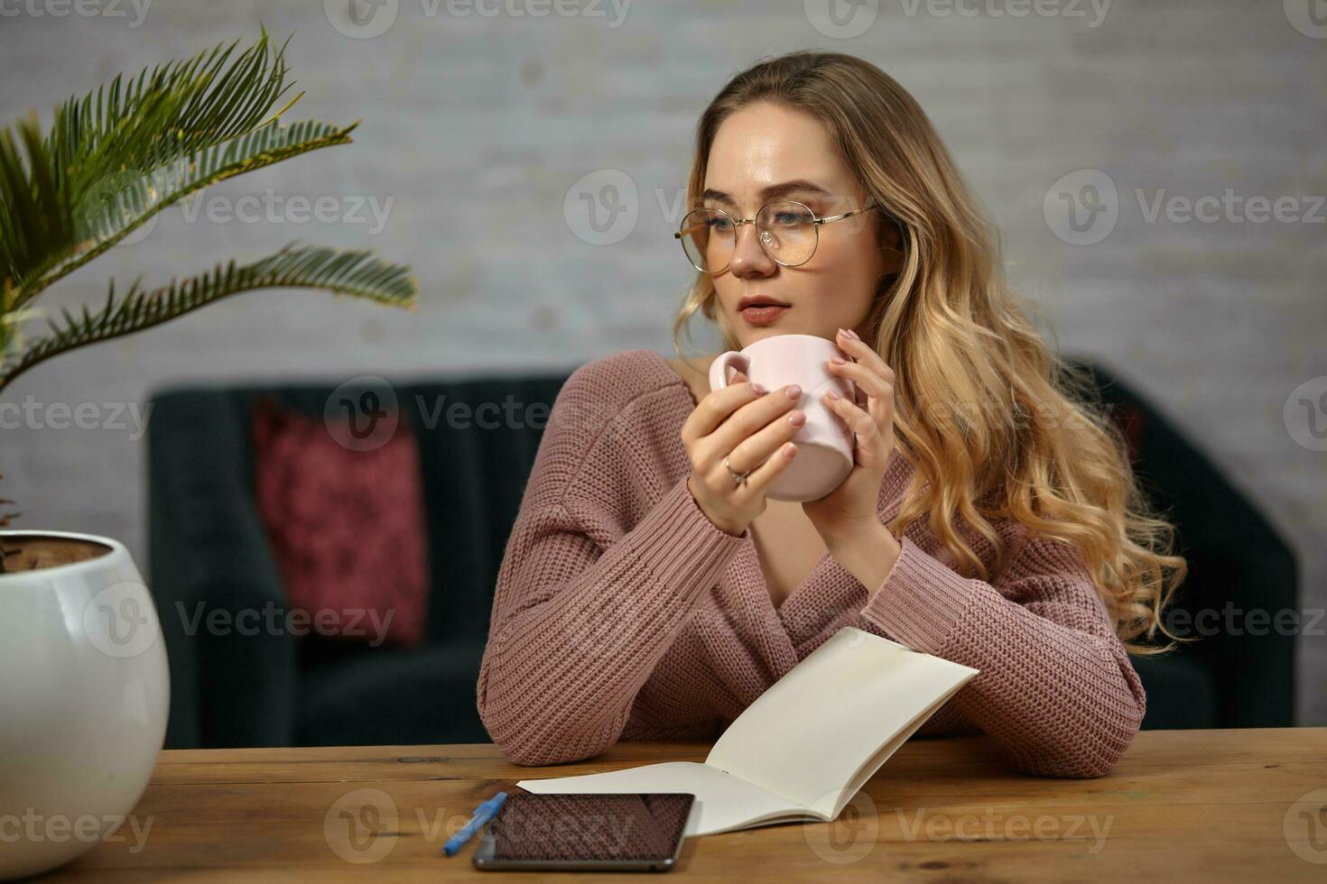 Female in glasses, pink cardigan. Holding a cup, sitting at wooden table with a tablet, notebook and flower in pot on it. Student, blogger. Close-up photo