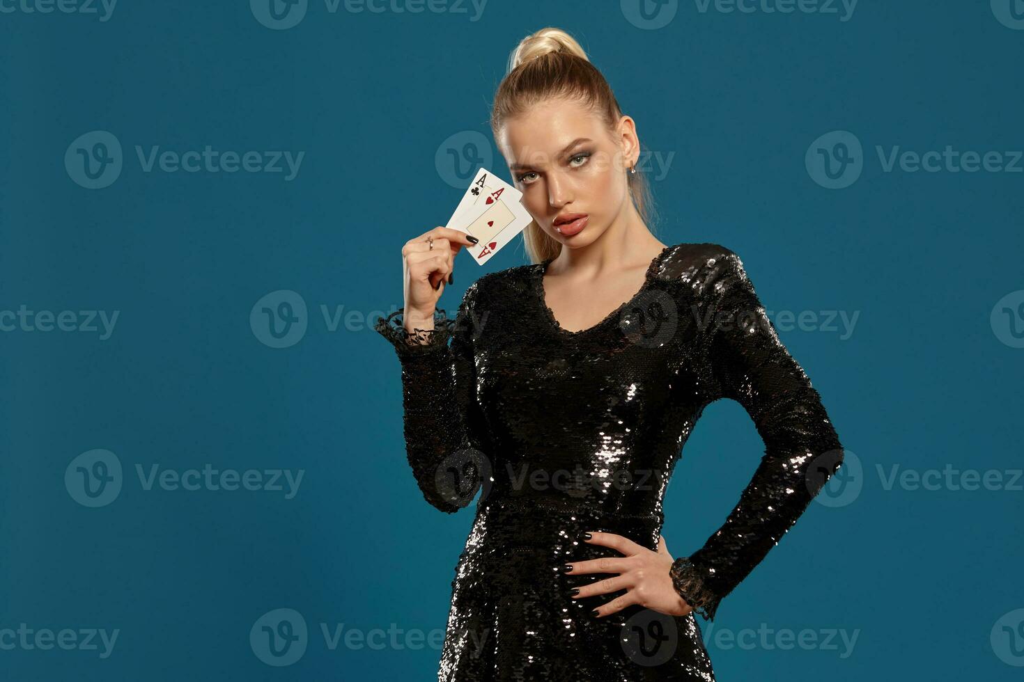 Blonde girl in black sequin dress showing two playing cards, posing against blue background. Gambling entertainment, poker, casino. Close-up photo
