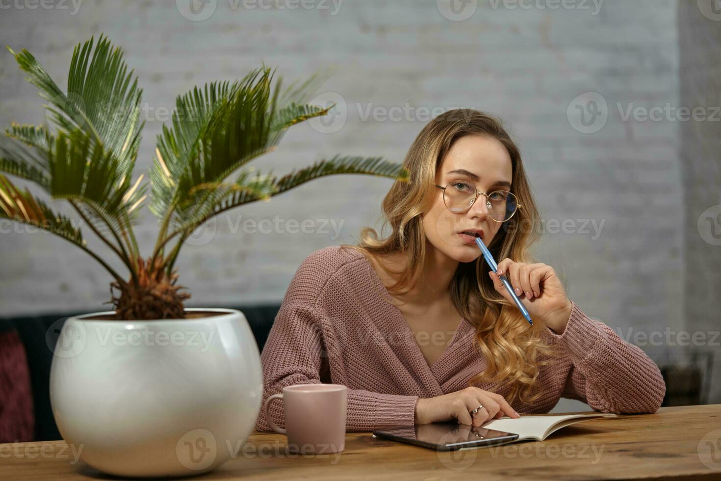Girl blogger in pink cardigan and glasses. Sitting at wooden table, going to write something in notebook. Palm tree in pot, cup and tablet nearby photo