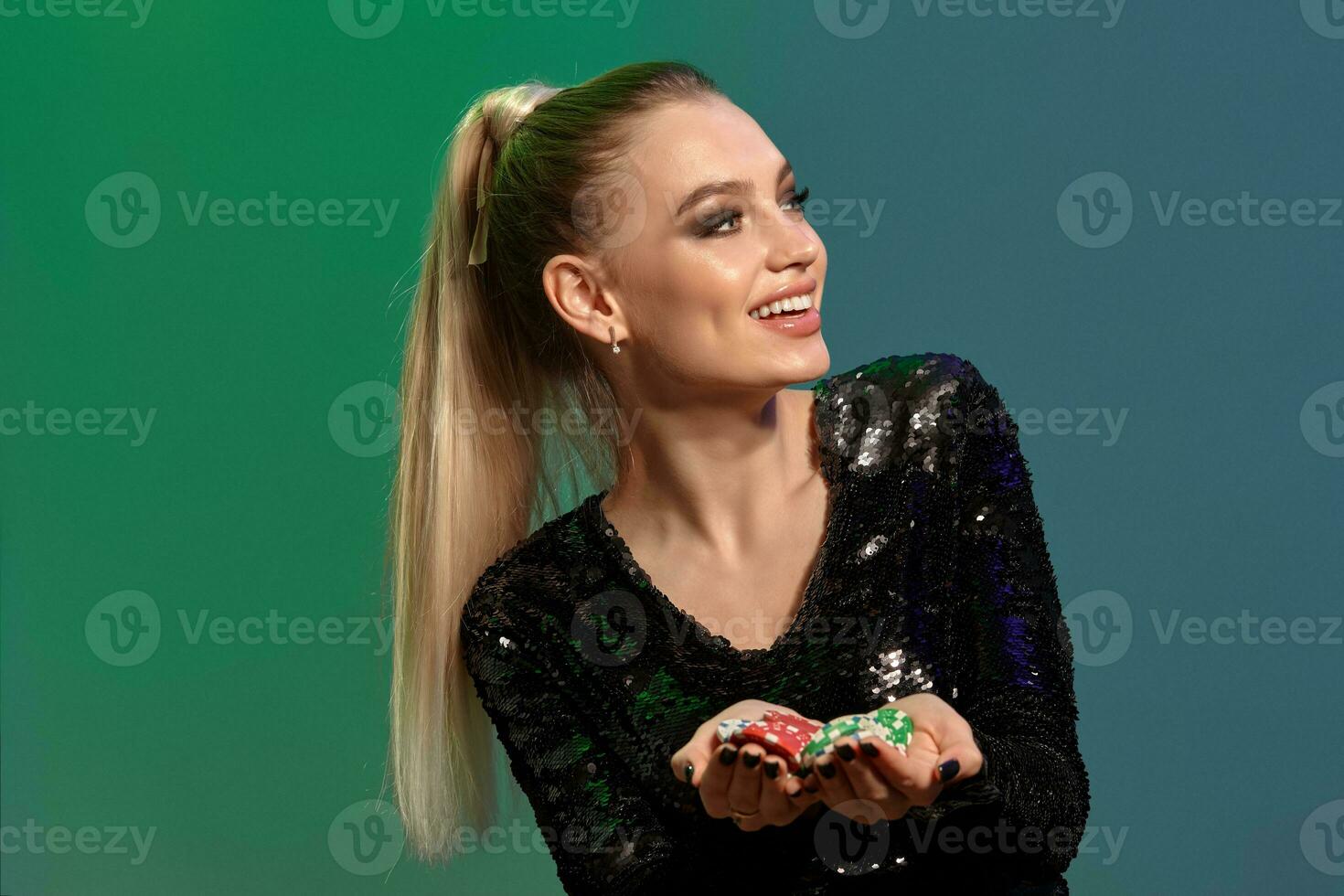 Blonde model in jewelry and black sequin dress. Smiling, showing handful of chips, posing on colorful background. Gambling, poker, casino. Close-up photo