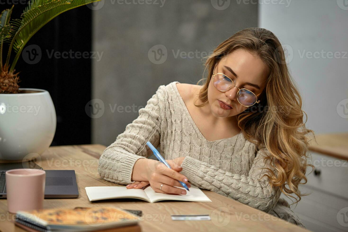 Blonde model blogger in glasses, beige sweater. Sitting in kitchen at wooden table, going to write something in her notebook. Close up photo