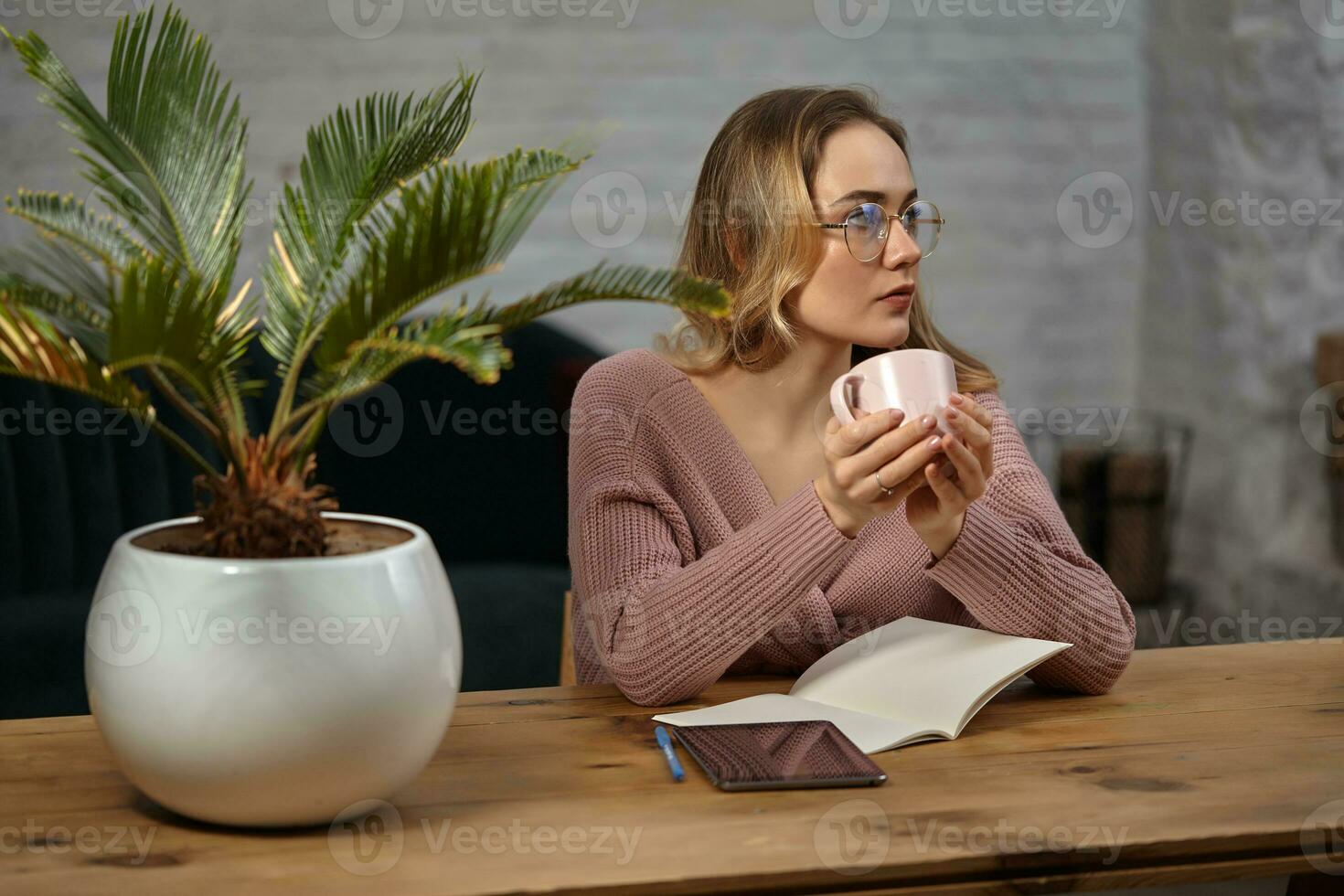 Woman glasses, pink cardigan. Holding a cup, sitting at wooden table with a tablet, notebook and flower in pot on it. Student, blogger. Close-up photo