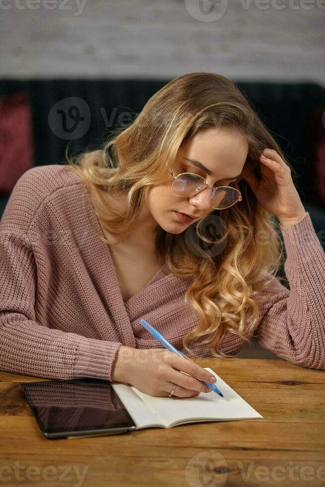 Blonde lady blogger in pink cardigan and glasses. Sitting at wooden table, going to write something in her notebook. Tablet is lying nearby. Close up photo