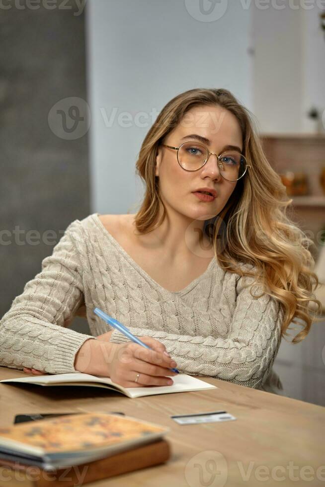 Blonde female blogger in glasses, beige sweater. Sitting in kitchen at wooden table, going to write something in her notebook. Close up photo