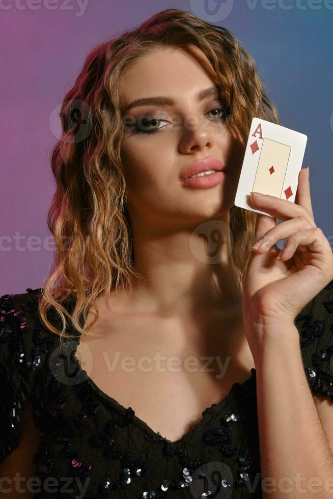 Brown-haired lady in black shiny dress showing ace of diamonds, posing on colorful background. Gambling entertainment, poker, casino. Close-up. photo