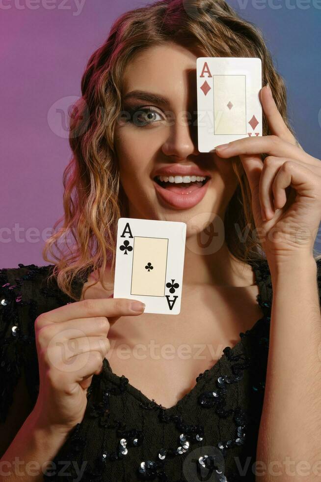 Brown-haired female in black shiny dress is smiling, showing two playing cards, posing on colorful background. Gambling, poker, casino. Close-up. photo