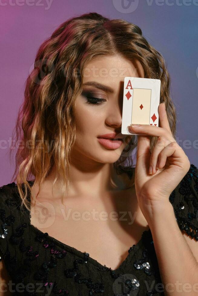 Brown-haired girl in black shiny dress has covered her eye with ace of diamonds, posing on colorful background. Gambling, poker, casino. Close-up. photo