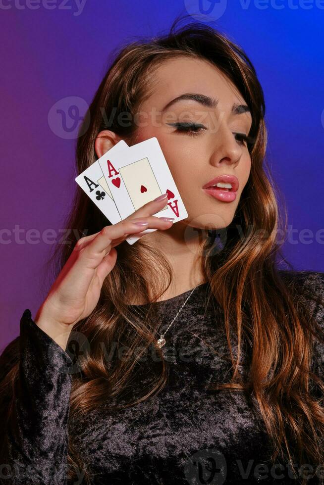 Brunette woman in black velvet dress showing two playing cards, posing against coloful background. Gambling entertainment, poker, casino. Close-up. photo