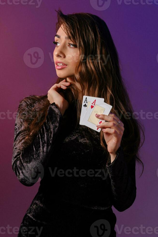 morena mujer en negro terciopelo vestir demostración dos jugando tarjetas, posando en contra coloful antecedentes. juego entretenimiento, póker, casino. de cerca. foto