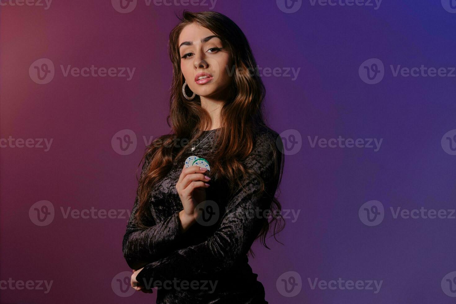 Brunette woman in black velvet dress showing multicolored chips, posing against coloful background. Gambling entertainment, poker, casino. Close-up. photo