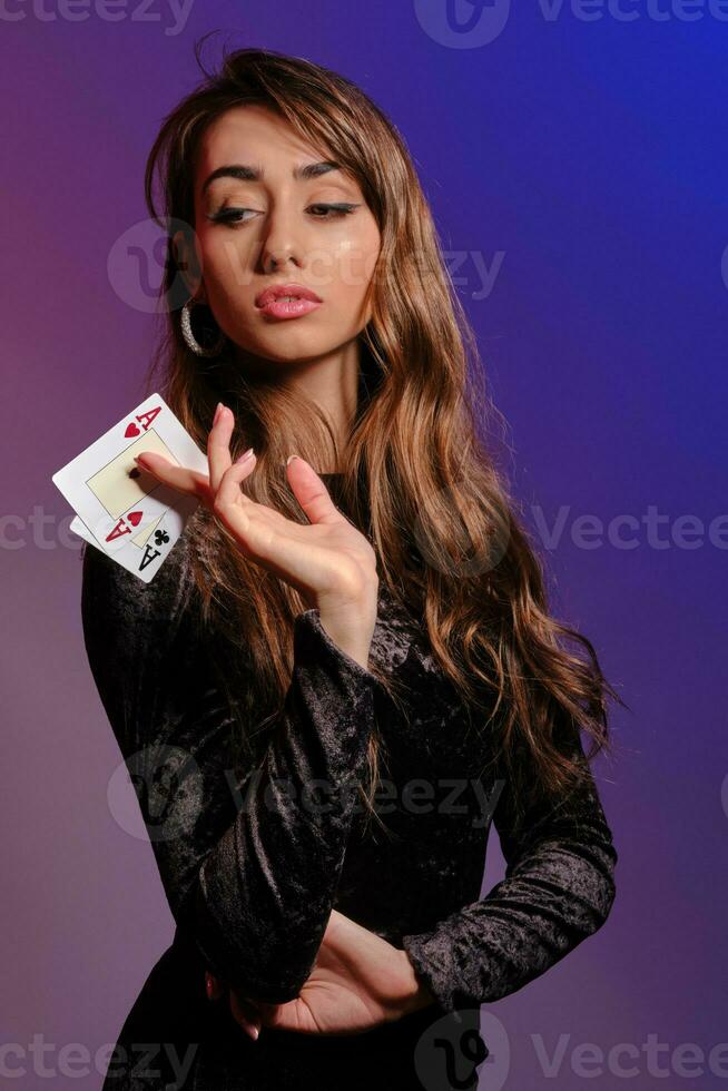 Brunette woman in black velvet dress showing two playing cards, posing against coloful background. Gambling entertainment, poker, casino. Close-up. photo