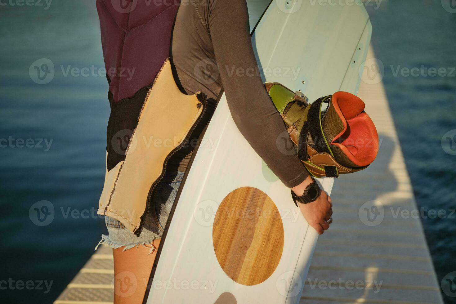 Brunette girl in a gray turtleneck, special sports vest and blue denim shorts is posing with her wakeboard on a pier of the coastal zone. Close-up. photo