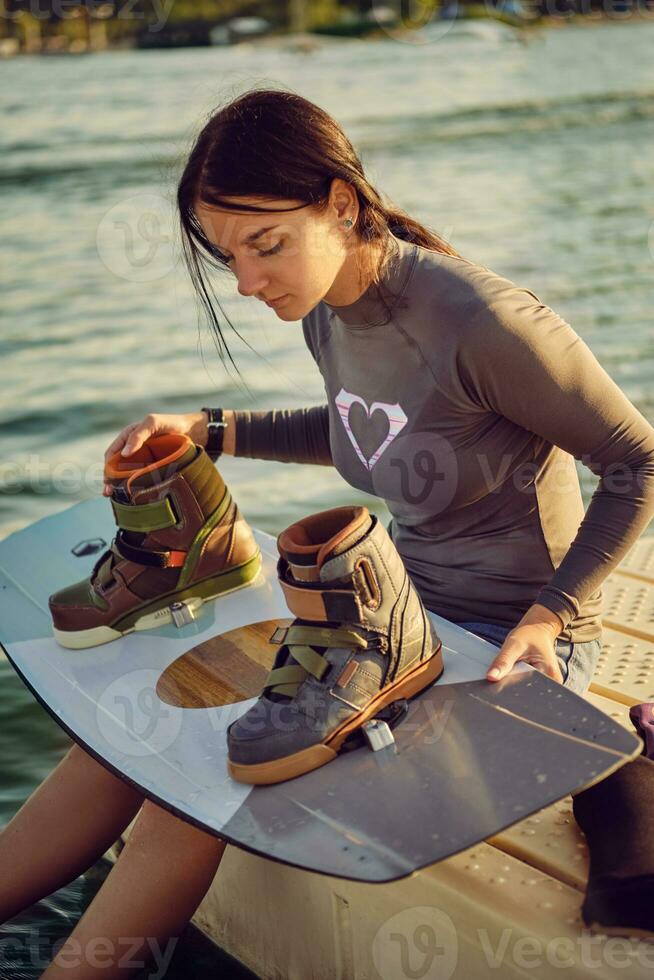 Brunette girl in a gray turtleneck and blue denim shorts is checking up boots on her wakeboard while sitting on a pier of the coastal zone. Close-up. photo