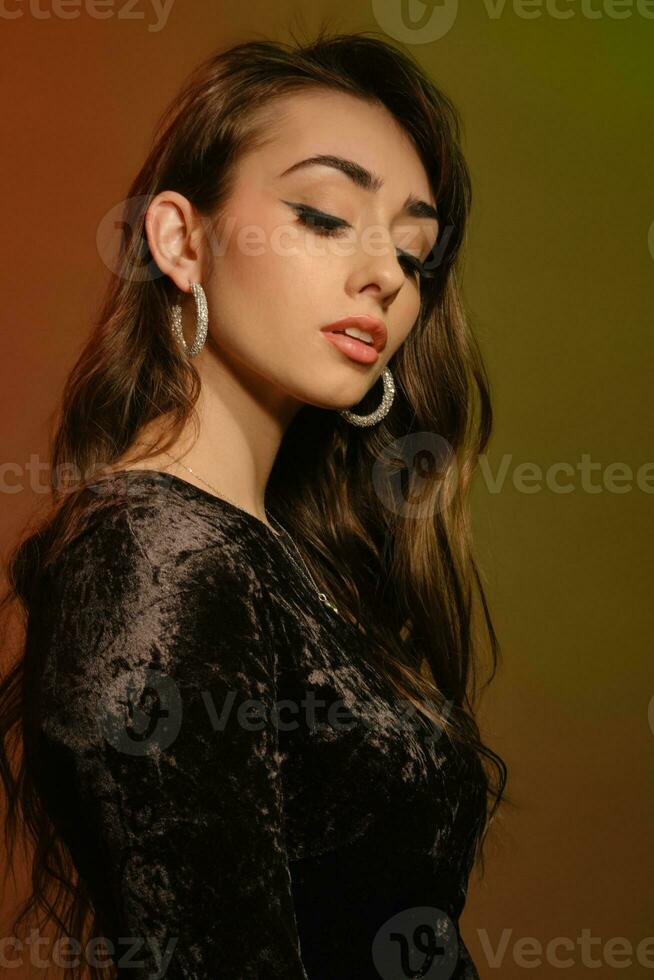 Attractive brunette woman in stylish jewelry and black velvet dress, posing sideways on colorful background. Fashion and beauty. Close-up, copy space photo