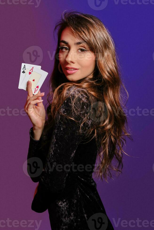 Brunette girl in black velvet dress showing two playing cards, posing sideways on coloful background. Gambling entertainment, poker, casino. Close-up. photo