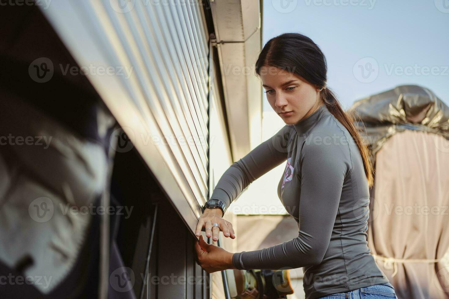 Beautiful brunette female in gray turtleneck and blue denim shorts is closing her wakeboard and equipment storage garage. Close-up. photo