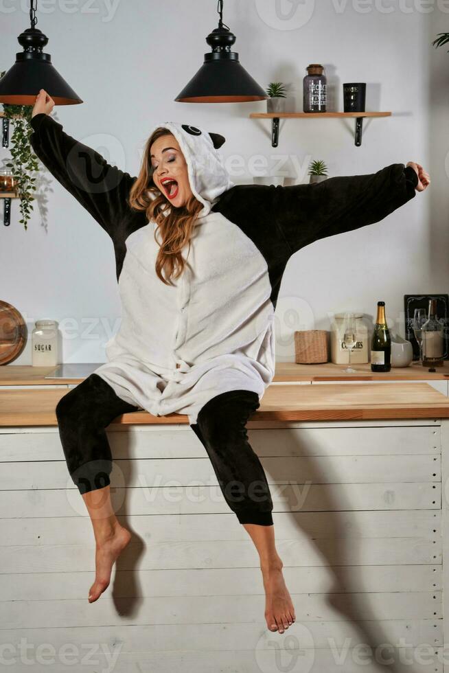 Blonde woman in plush pajama in form of cartoon character panda is having fun at bachelorette party. Yawning, sitting on kitchen table. Close-up. photo